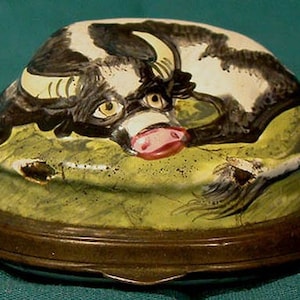 May include: A vintage porcelain trinket box with a hand-painted design of a black and white cow lying down on a green field. The box has a brass base.