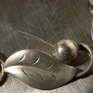 K&ouml;nnte beinhalten: Ein silbernes Armband mit blattf&ouml;rmigen Anh&auml;ngern und runden Perlen. Die Bl&auml;tter haben eine strukturierte Oberfl&auml;che und sind durch silberne Ringe miteinander verbunden.