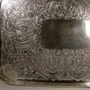 May include: A silver serving tray with an ornate, embossed design. The tray is rectangular in shape and has a raised rim.