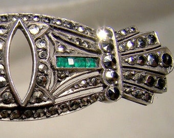 May include: A silver brooch with a delicate design featuring a central oval opening and two emerald green stones. The brooch is adorned with numerous small, sparkling stones.