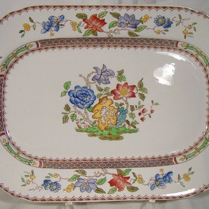 May include: A white oval serving platter with a decorative floral design. The border features a repeating pattern of brown, red, and green. The center of the platter features a cluster of blue, red, and yellow flowers with green leaves.
