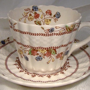 May include: A white teacup and saucer with a brown and floral pattern. The cup has a scalloped rim and a handle. The saucer has a scalloped edge.