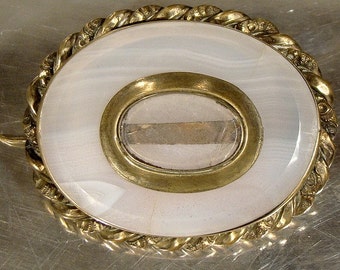 May include: A white cameo brooch with a gold-toned frame and a decorative rope-like border. The cameo is set in a gold-toned oval frame with a glass window.
