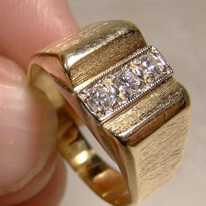 May include: A gold ring with three small diamonds set in a row. The ring has a brushed finish and a square-shaped band.