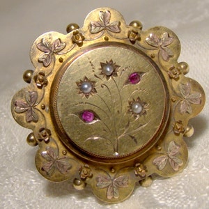 May include: A gold-toned brooch with a circular design featuring a floral motif with pink and red stones. The brooch is surrounded by a scalloped edge with shamrock designs.