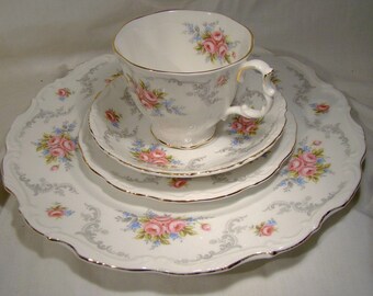 May include: A white teacup and saucer set with a delicate floral pattern featuring pink roses and blue forget-me-nots. The set is trimmed with a gold band and has a scalloped edge.
