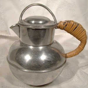 May include: A silver pitcher with a woven brown handle. The pitcher has a rounded body and a lid.