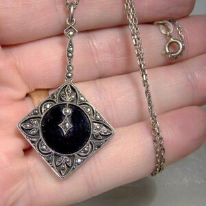 May include: A silver necklace with a black onyx pendant. The pendant is square shaped and features a detailed design of marcasite stones. The chain is a delicate silver chain.