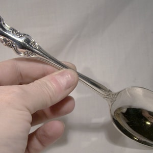 May include: A silver spoon with an ornate handle and a rounded bowl. The spoon is held in a hand.