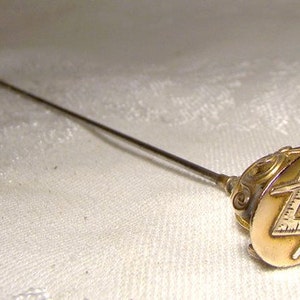 May include: A gold-toned hat pin with a Masonic symbol on the top. The symbol is a compass and square with a letter 'G' in the center.