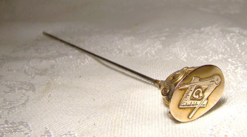 May include: A gold-toned hat pin with a Masonic symbol on the top. The symbol is a compass and square with a letter 'G' in the center.