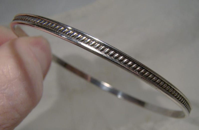 K&ouml;nnte beinhalten: Ein silbernes Armreif-Armband mit einem strukturierten Seildesign an den R&auml;ndern. Das Armband ist rund und scheint aus Metall zu sein. Der Armreif ist ein schlichtes, elegantes Schmuckst&uuml;ck.