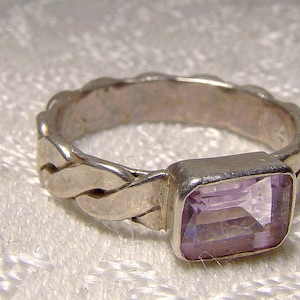 May include: A silver ring with a braided band and a light purple rectangular gemstone.