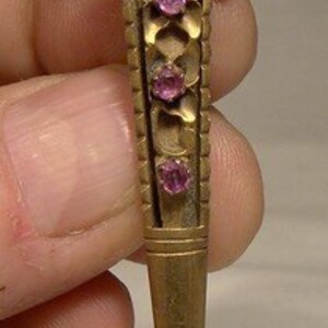 May include: A gold-toned brooch with a tree-like design and pink rhinestones. The brooch is shaped like a long, thin pin.