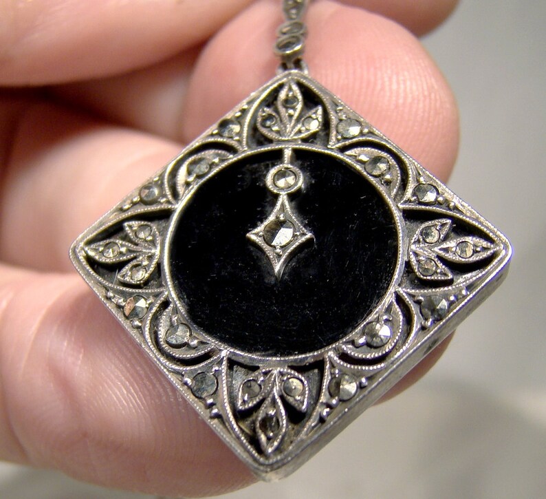 May include: A silver pendant with a black enamel center and a geometric design. The pendant is decorated with marcasite stones.