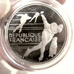 May include: Silver coin featuring a stylized image of a speed skater, a groundhog, and the text "REPUBLIQUE FRANCAISE".