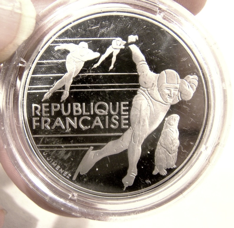May include: Silver coin featuring a stylized image of a speed skater, a groundhog, and the text "REPUBLIQUE FRANCAISE".