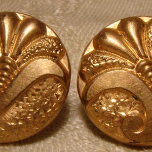 May include: Two antique gold-colored buttons with an ornate, Art Deco-inspired design. Each button features a central, stylized floral element and swirling, textured patterns. The buttons are round and have a slightly raised relief.