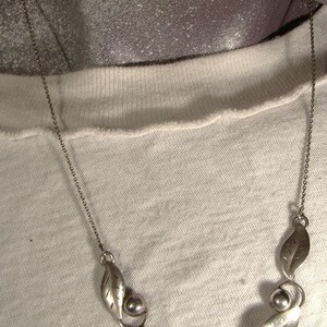 K&ouml;nnte beinhalten: Eine silberne Halskette mit einer zarten Kette und einem Design aus Bl&auml;ttern und Perlen. Die Halskette liegt auf einem wei&szlig;en Hintergrund.