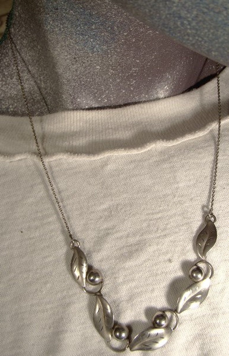 K&ouml;nnte beinhalten: Eine silberne Halskette mit einer zarten Kette und einem Design aus Bl&auml;ttern und Perlen. Die Halskette liegt auf einem wei&szlig;en Hintergrund.