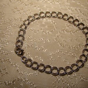 May include: Silver bracelet with a double-link chain design. The bracelet is secured with a small clasp. The jewellery is displayed on a textured, light-coloured surface.
