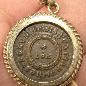 May include: A gold-toned pendant with a rope-like border. The pendant features a round, silver-colored coin with the inscription "IMPERATOR CAESAR DIVI AVG F AVGVSTVS" and "IOA" in the centre.