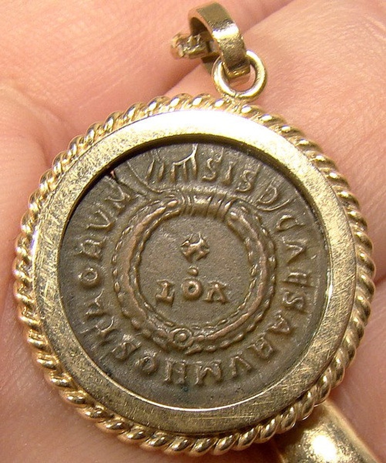May include: A gold-toned pendant with a rope-like border. The pendant features a round, silver-colored coin with the inscription "IMPERATOR CAESAR DIVI AVG F AVGVSTVS" and "IOA" in the centre.