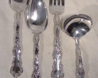 May include: Four pieces of ornate silver flatware, including two spoons, a fork, and a ladle. The silverware has detailed, decorative handles and a polished, reflective surface. The pieces are arranged on a light-colored surface.