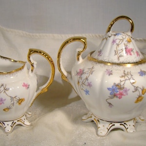 May include: A white porcelain creamer and sugar bowl set with gold trim and floral designs. The creamer and sugar bowl have a scalloped edge and are decorated with pink, blue, and yellow flowers.