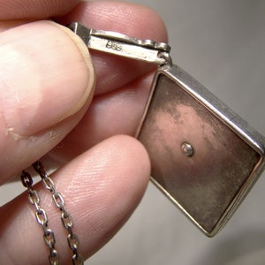 May include: Silver pendant with a square-shaped charm. The charm has a smooth, silver surface with a small, round, clear stone in the center. The pendant is attached to a silver chain. The back of the pendant is marked with the number "925".