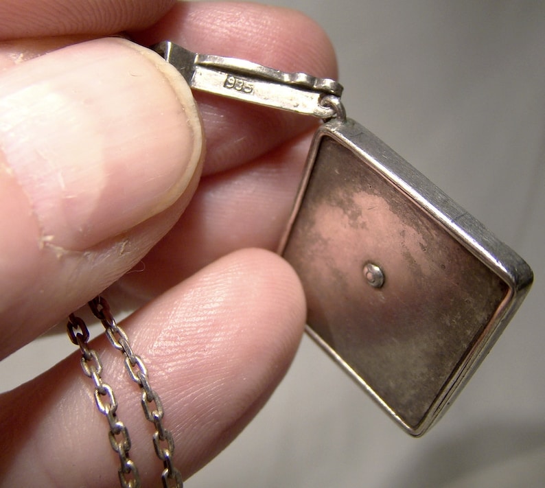 May include: Silver pendant with a square-shaped charm. The charm has a smooth, silver surface with a small, round, clear stone in the center. The pendant is attached to a silver chain. The back of the pendant is marked with the number "925".