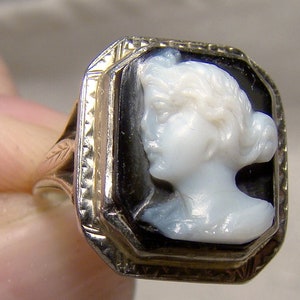 May include: A gold ring with a black and white cameo featuring a woman's profile. The cameo is set in a square frame with a decorative border.