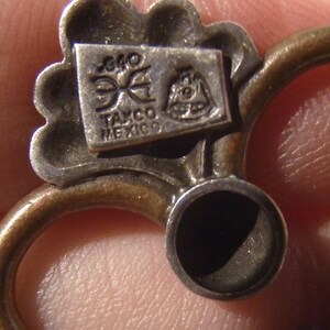 May include: Close-up of a silver charm with a floral design. The charm has a small rectangular tag with the text "Taxco Mexico" and a logo.