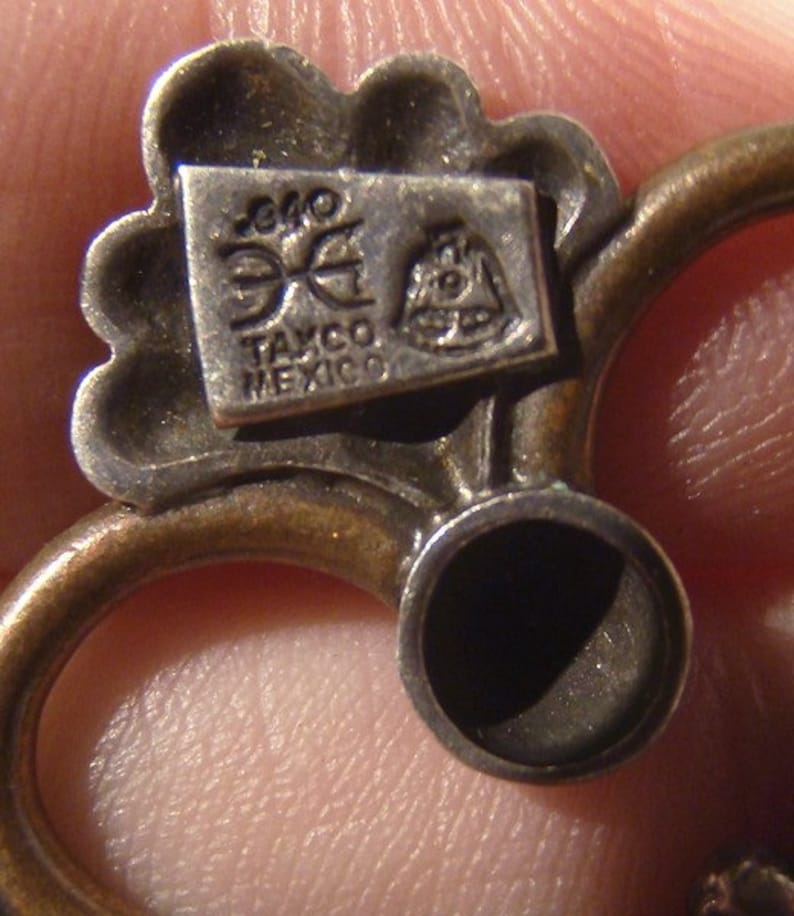 May include: Close-up of a silver charm with a floral design. The charm has a small rectangular tag with the text "Taxco Mexico" and a logo.