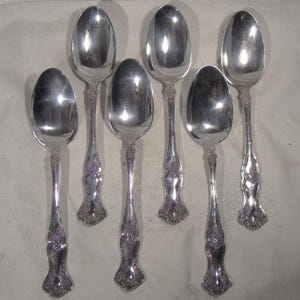 May include: Six silver spoons with ornate floral detailing on the handles. The spoons have a classic, elegant design, with a polished, reflective surface. The spoons are arranged on a light-colored surface, suggesting a formal dining setting.
