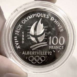 May include: Silver coin commemorating the XVI Olympic Winter Games in Albertville, France in 1992. The coin features the Olympic rings, a stylized flame, and the text "XVIes Jeux Olympiques d'Hiver 1992 Albertville 92 100 Francs".