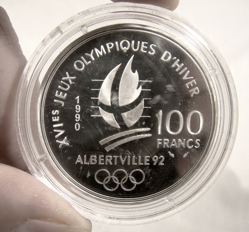 May include: Silver coin commemorating the XVI Olympic Winter Games in Albertville, France in 1992. The coin features the Olympic rings, a stylized flame, and the text "XVIes Jeux Olympiques d'Hiver 1992 Albertville 92 100 Francs".