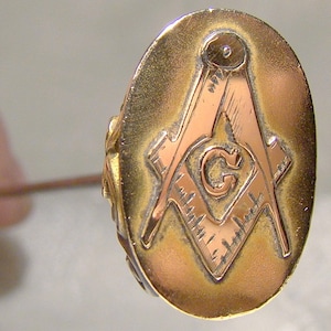 May include: A gold-toned oval pin with a copper-colored Masonic symbol in the center. The symbol is a square and compass with a 'G' in the center.