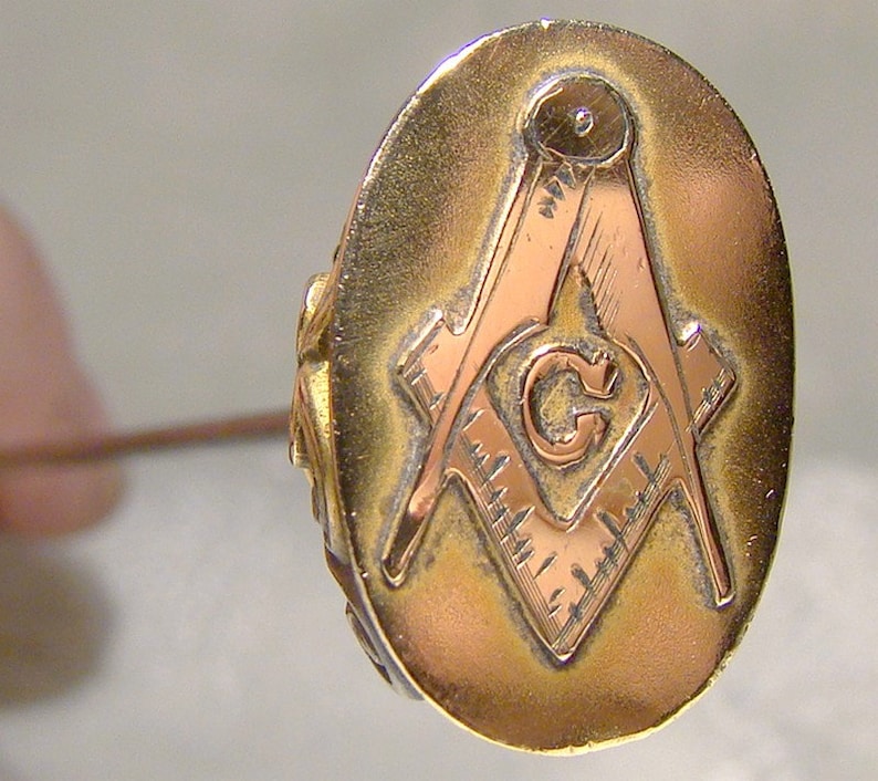 May include: A gold-toned oval pin with a copper-colored Masonic symbol in the center. The symbol is a square and compass with a 'G' in the center.