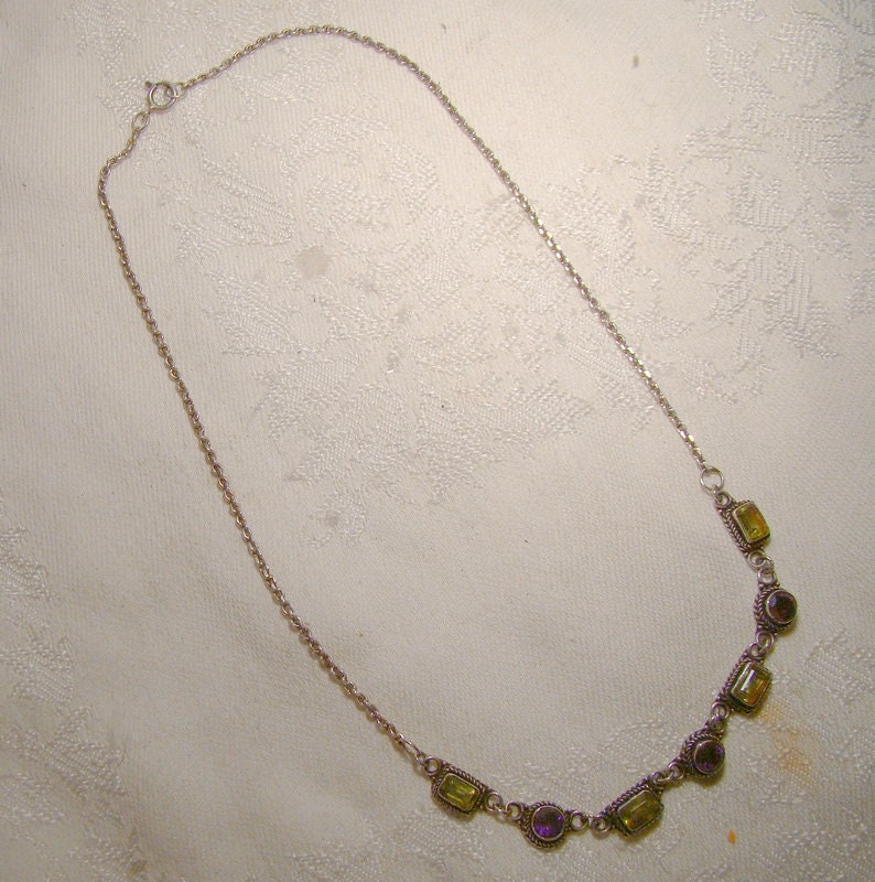 May include: A silver chain necklace with a row of five rectangular green and purple gemstones.