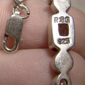 Puede incluir: Un brazalete de cadena de plata con un dije rectangular que dice "R8G 925".
