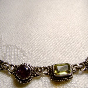 May include: A silver necklace featuring alternating green and purple gemstones. The necklace is made from a delicate chain and has a simple design.