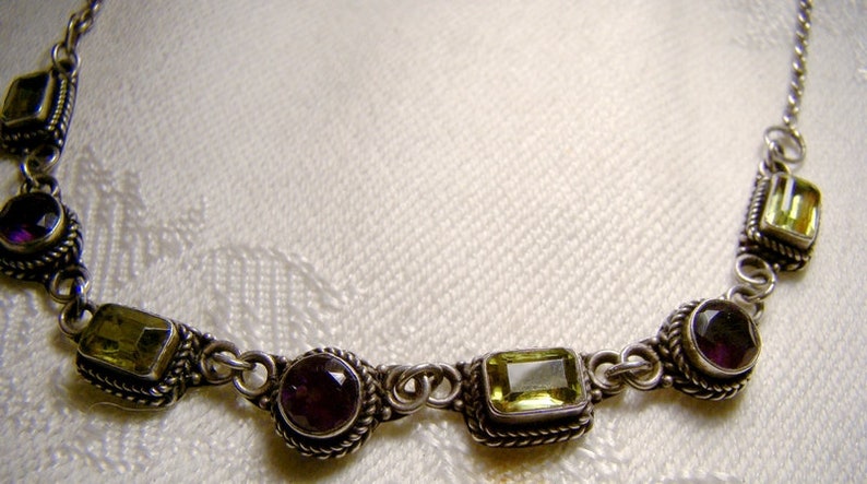 May include: A silver necklace featuring alternating green and purple gemstones. The necklace is made from a delicate chain and has a simple design.