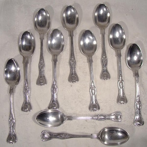 May include: A collection of nine silver spoons with ornate handles and oval bowls. The spoons are arranged on a light-colored surface, showcasing their reflective qualities and intricate designs. The spoons vary slightly in size and shape.