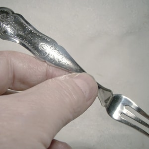 May include: A silver fork with an ornate handle and three tines. The handle is decorated with a floral design.