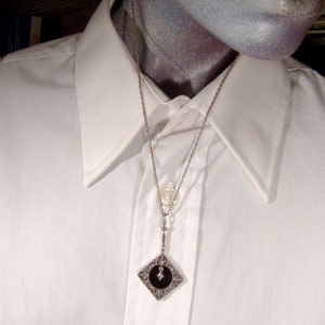 May include: A silver chain necklace with a black and silver pendant. The pendant is a square with a black center and silver filigree around the edges. The chain is delicate and has a small silver charm attached to it.