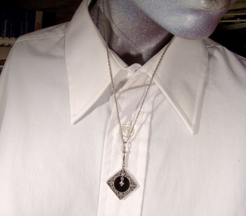 May include: A silver chain necklace with a black and silver pendant. The pendant is a square with a black center and silver filigree around the edges. The chain is delicate and has a small silver charm attached to it.