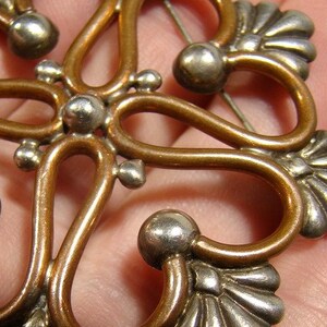 May include: A copper and silver brooch with a floral design. The brooch has a delicate, intricate design with rounded edges and a pin on the back.
