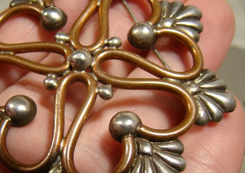 May include: A copper and silver brooch with a floral design. The brooch has a delicate, intricate design with rounded edges and a pin on the back.
