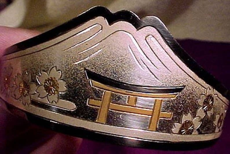 Pu&ograve; includere: Un braccialetto d'argento con una scena di montagna giapponese e un torii. Il braccialetto &egrave; decorato con fiori di ciliegio e ha un design in bianco e nero.
